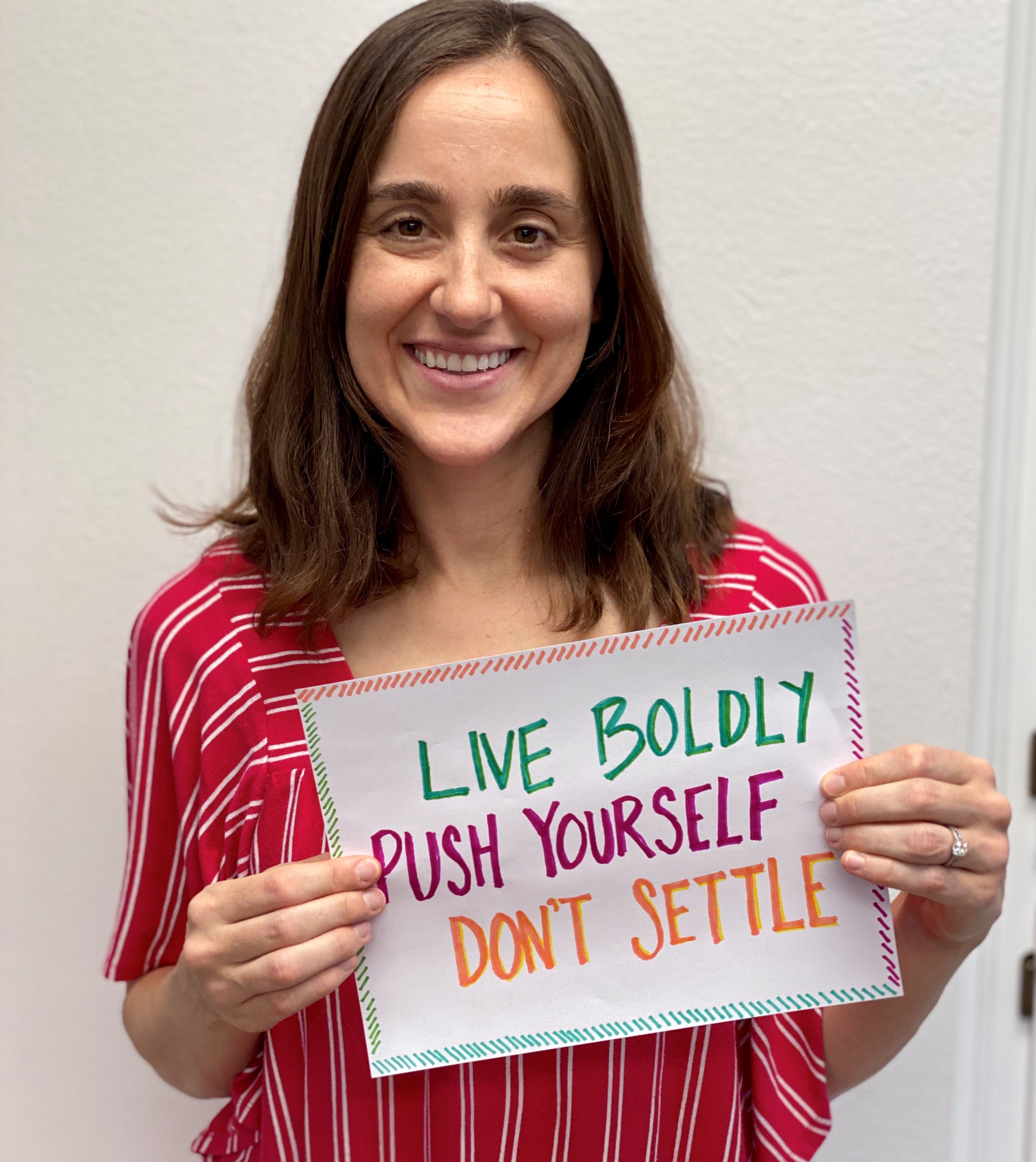 Brittany is smiling, holding a handmade sign which says "Live boldly, push yourself, don't settle."