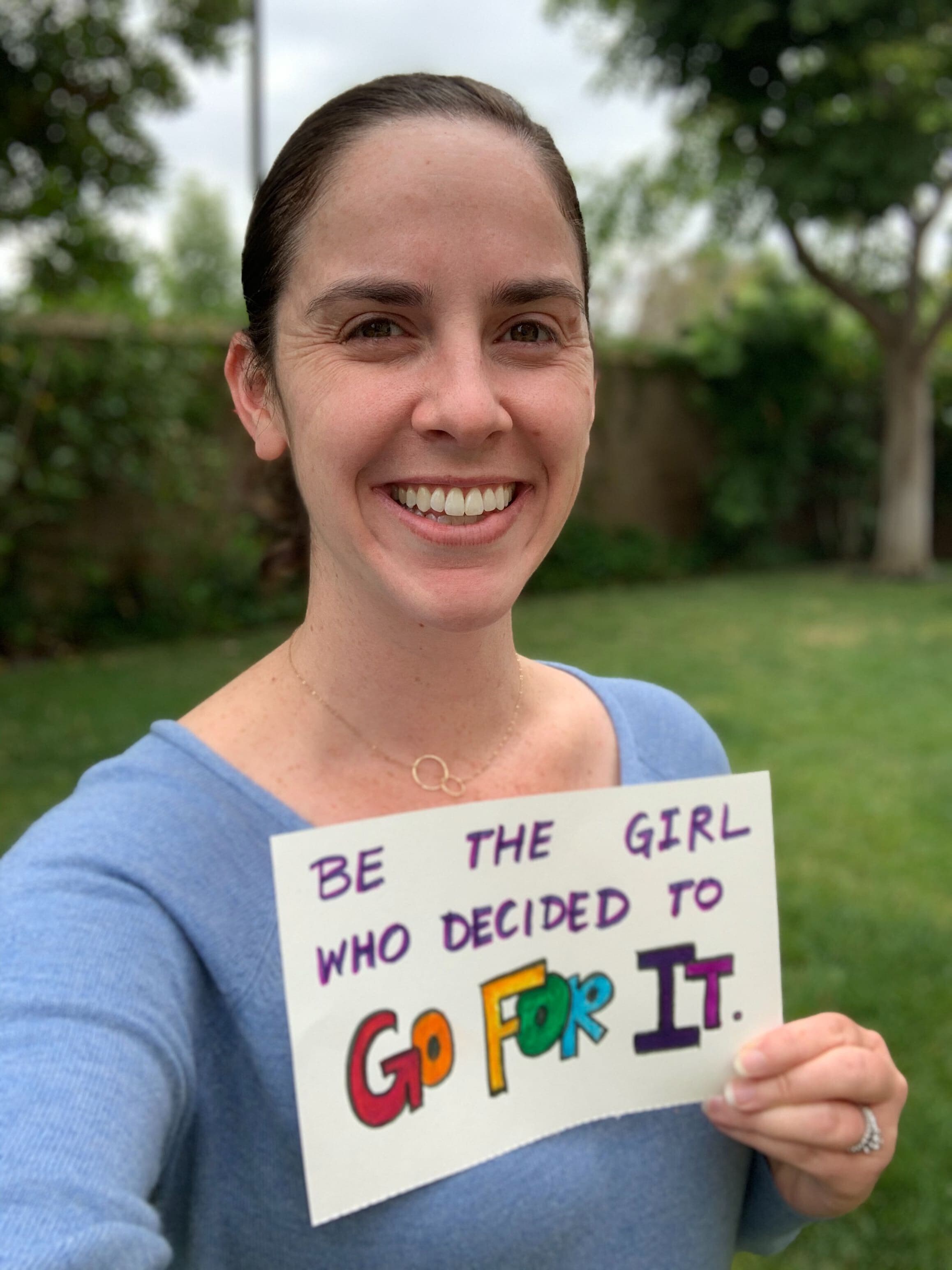 Rachelle is smiling, holding a handmade sign which says "Be the girl who decided to go for it!"