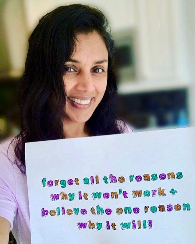 Kamini is smiling, holding a handmade sign which says "Forget the reasons why it won't work and believe the one reason why it will."